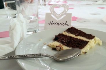 Ein Stück Hochzeittorte auf einem Teller. Links liegt ein Löffeln. Auf dem Tisch steht ein Namensschild, darauf steht "Heinrich".