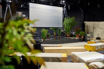 Eine große Leinwand hängt mitten im Raum. Darum herum stehen grüne Topfpflanzen. Vorn sieht man Kisten, auf denen man sitzen kann.