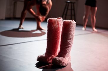 Große rosafarbene Pelzstiefel stehen auf einer dunklen Bühne. Im Hintergrund bewegen sich zwei Tänzer. Alles wirft scharfe Schatten.