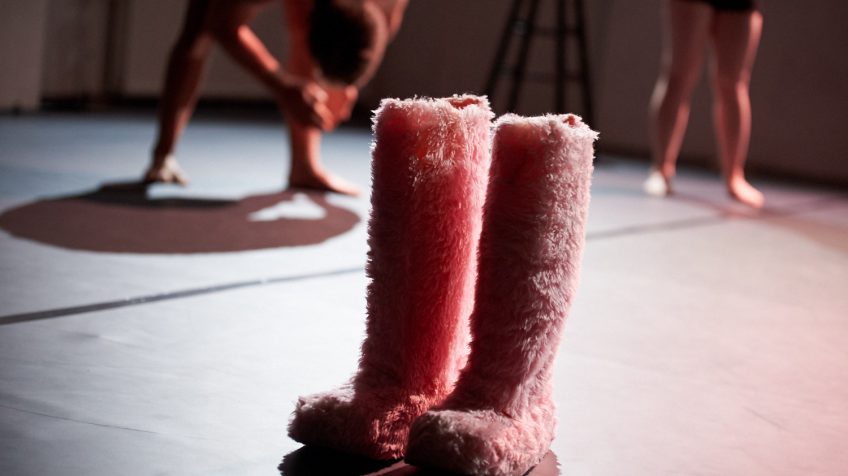 Große rosafarbene Pelzstiefel stehen auf einer dunklen Bühne. Im Hintergrund bewegen sich zwei Tänzer. Alles wirft scharfe Schatten.